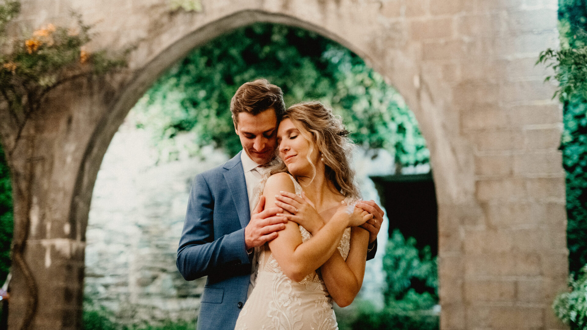 Groom embracing bride from behind with a stone archway in the background