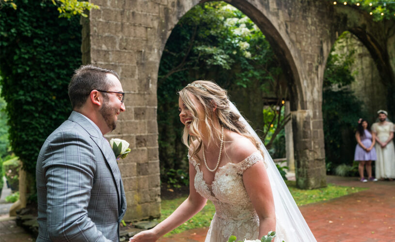 Bride and groom laughing outside on a patio with a stone arched wall in the background.