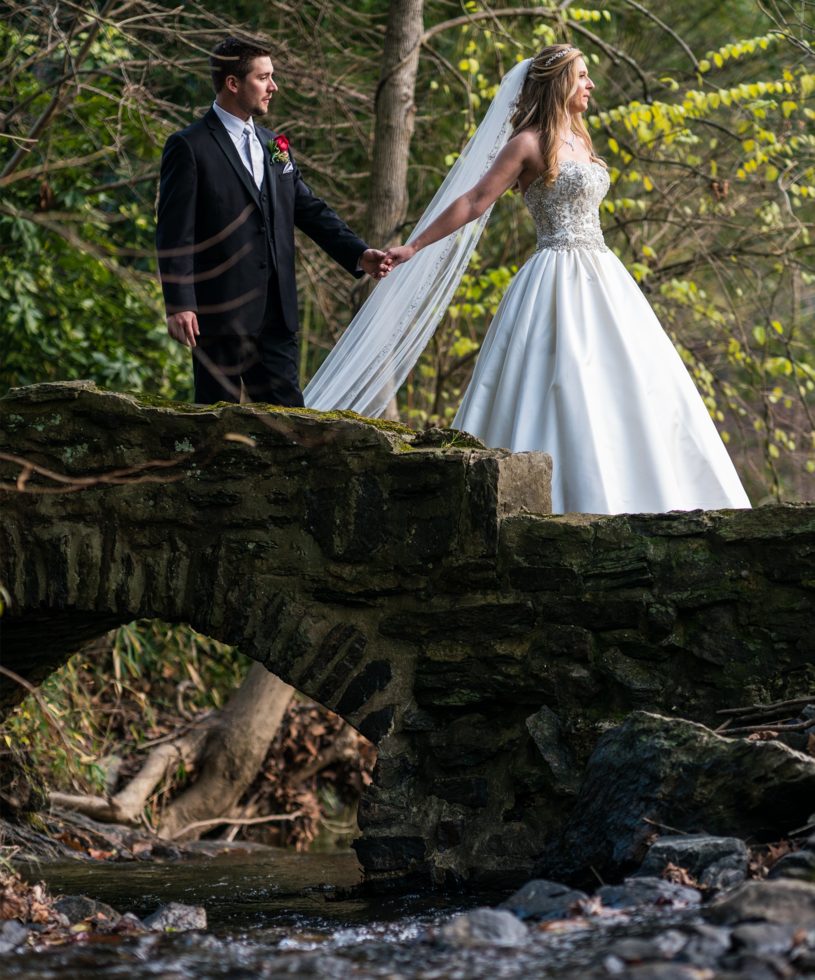 Bride and groom holding hands on stone bridge over small creek.