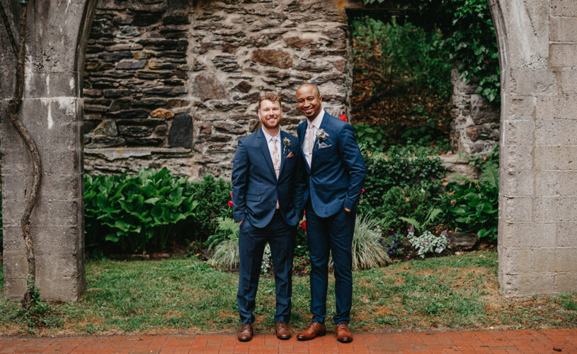 grooms standing outside under stone arch with stone wall and bushes behind them