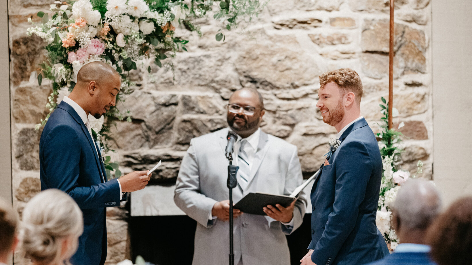 two grooms saying their vows in front of a preacher, flower archway, and stone wall