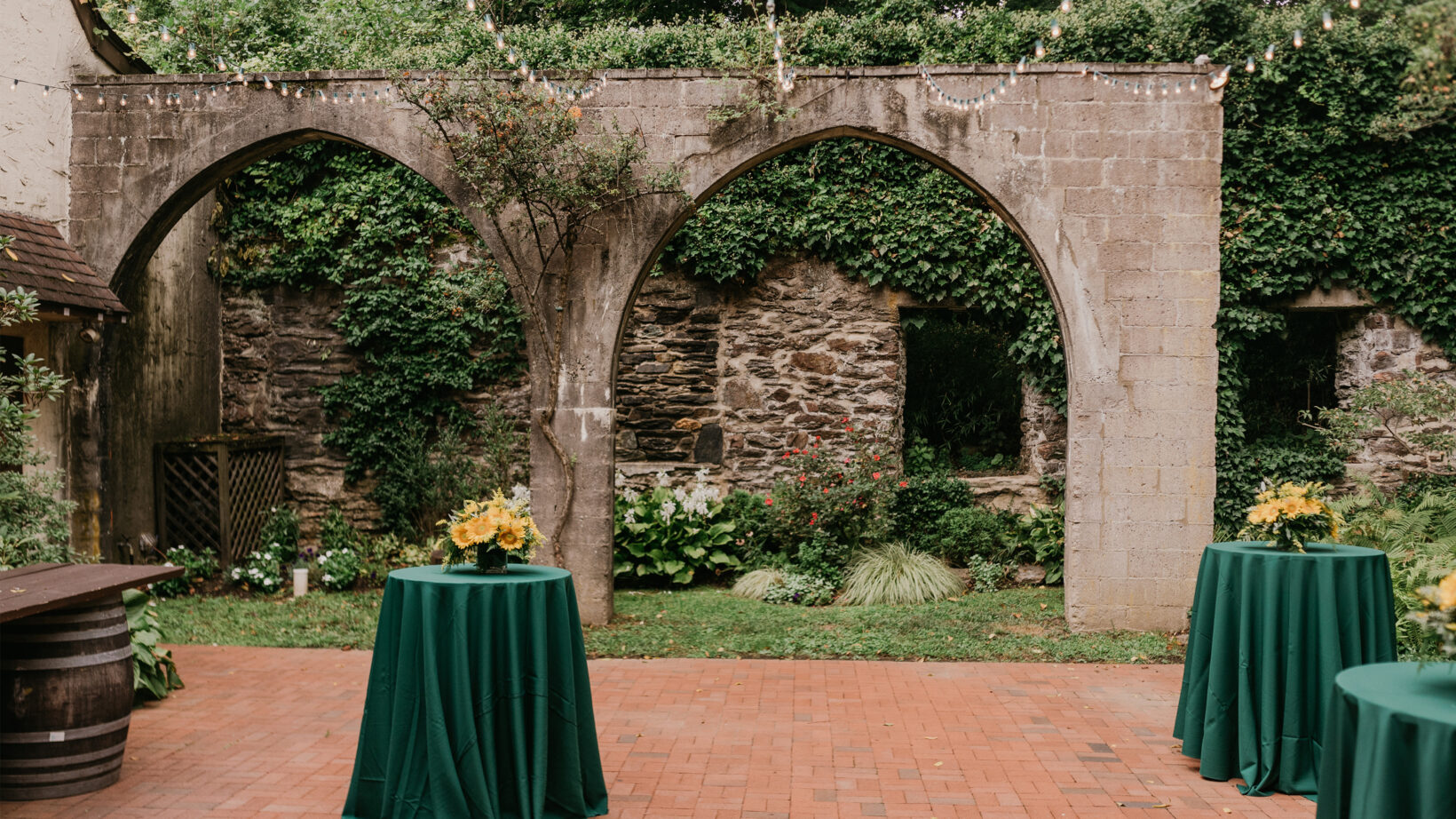 A few small round tables covered in a green table cloth outside on a stone patio next to stone archways